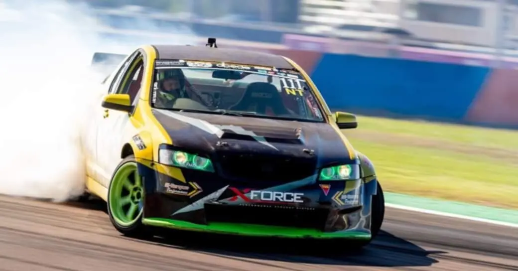 A race car with green wheels drifts on a track, emitting smoke. The vehicle has a black and yellow design with visible branding.