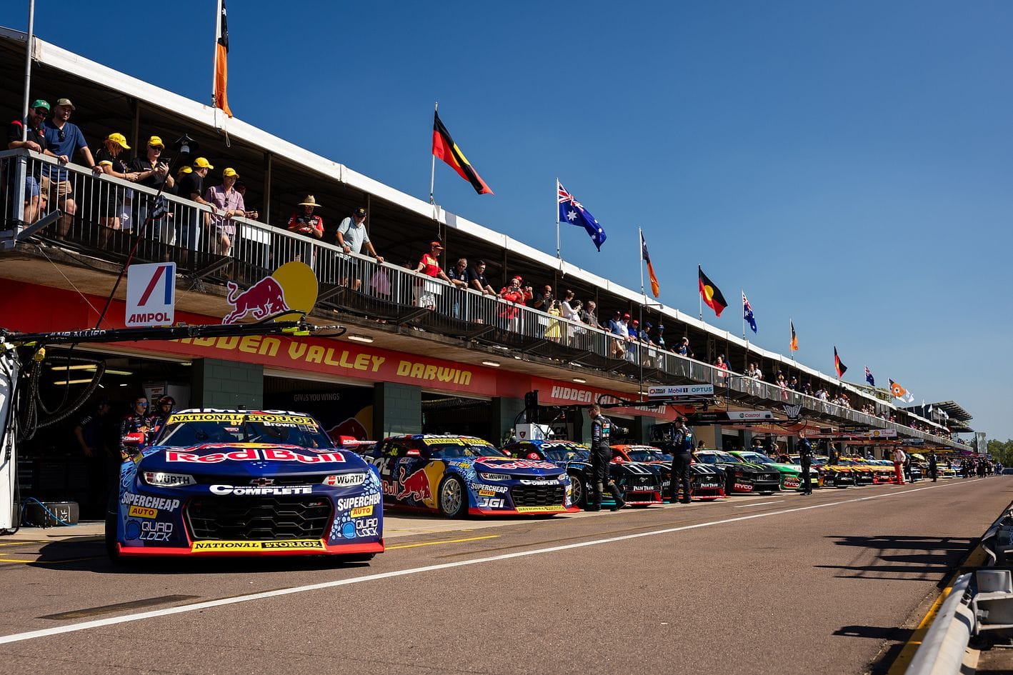 Race cars lined up in a pit lane under a clear blue sky, with spectators watching from a balcony above. Flags are displayed along the building.