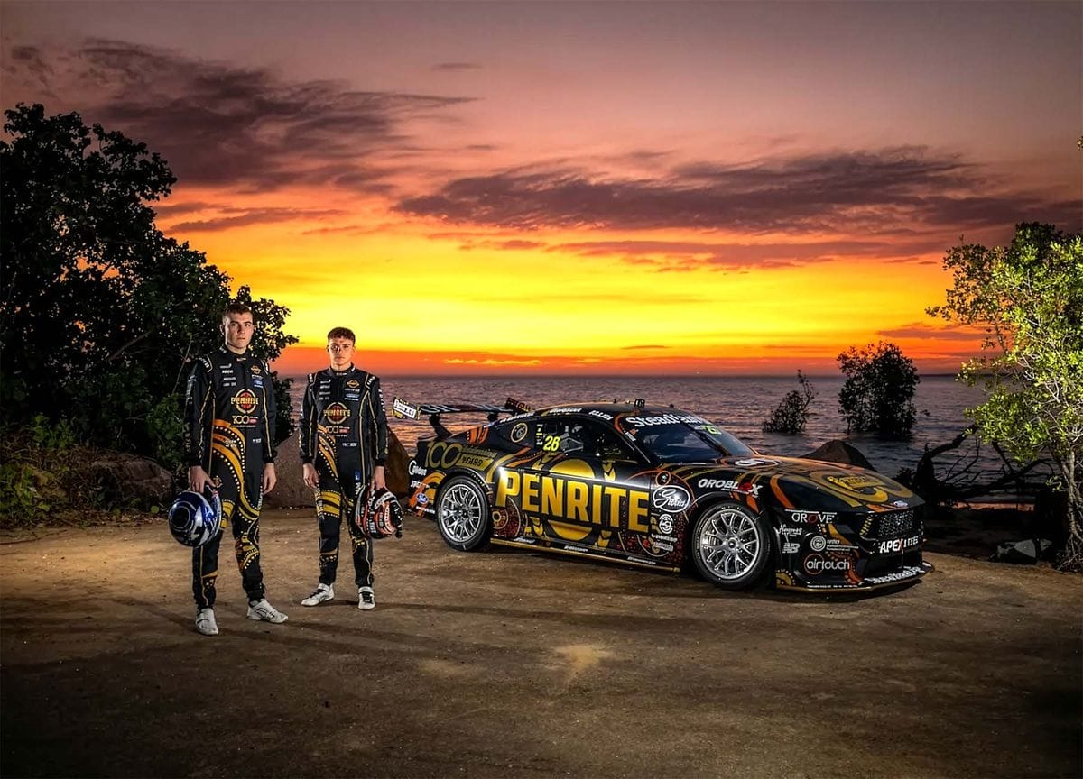 Two Penrite Racing drivers standing beside a black and gold Penrite Supercar at sunset, with the ocean in the background.