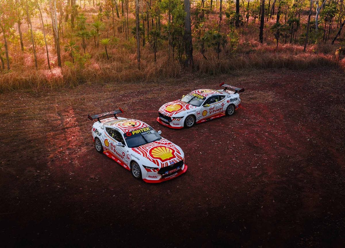 Two Shell V-Power Racing Team Supercars parked in bushland with warm sunlight through the trees.