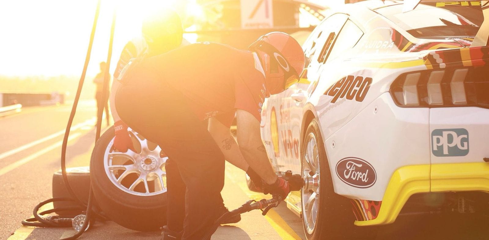 Mechanic in red gear changes a tire on a white race car at sunset in a pit stop, with sunlight creating a warm glow.