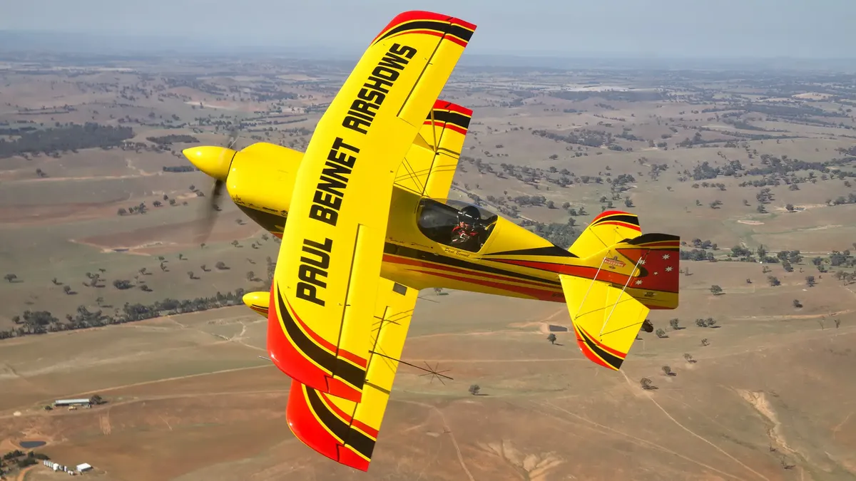A yellow biplane with "Paul Bennet Airshows" on the wings flies over a rural landscape, performing aerobatic maneuvers.
