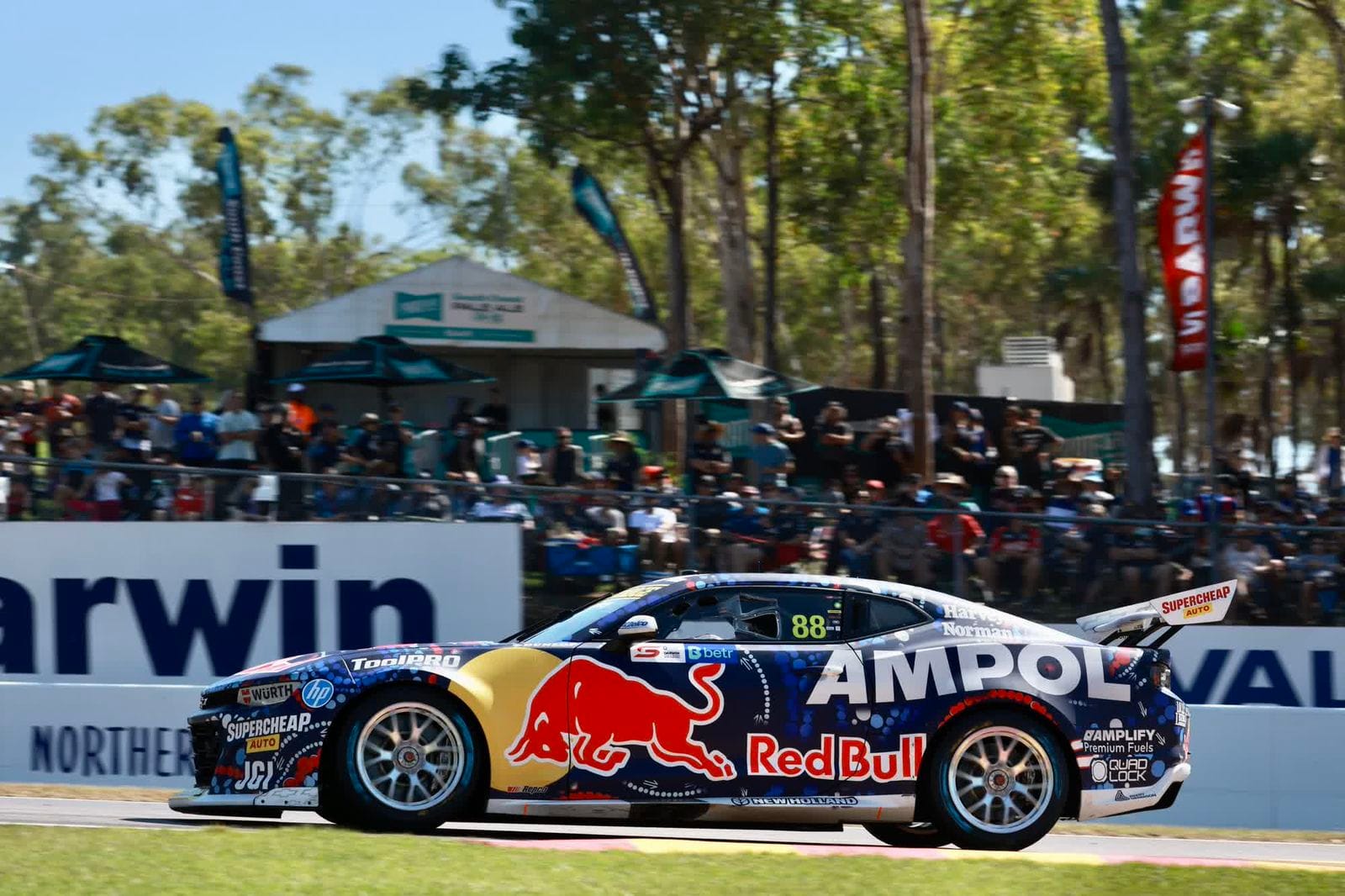 Race car with Red Bull and Ampol branding speeding on a track, surrounded by spectators and trees in the background.