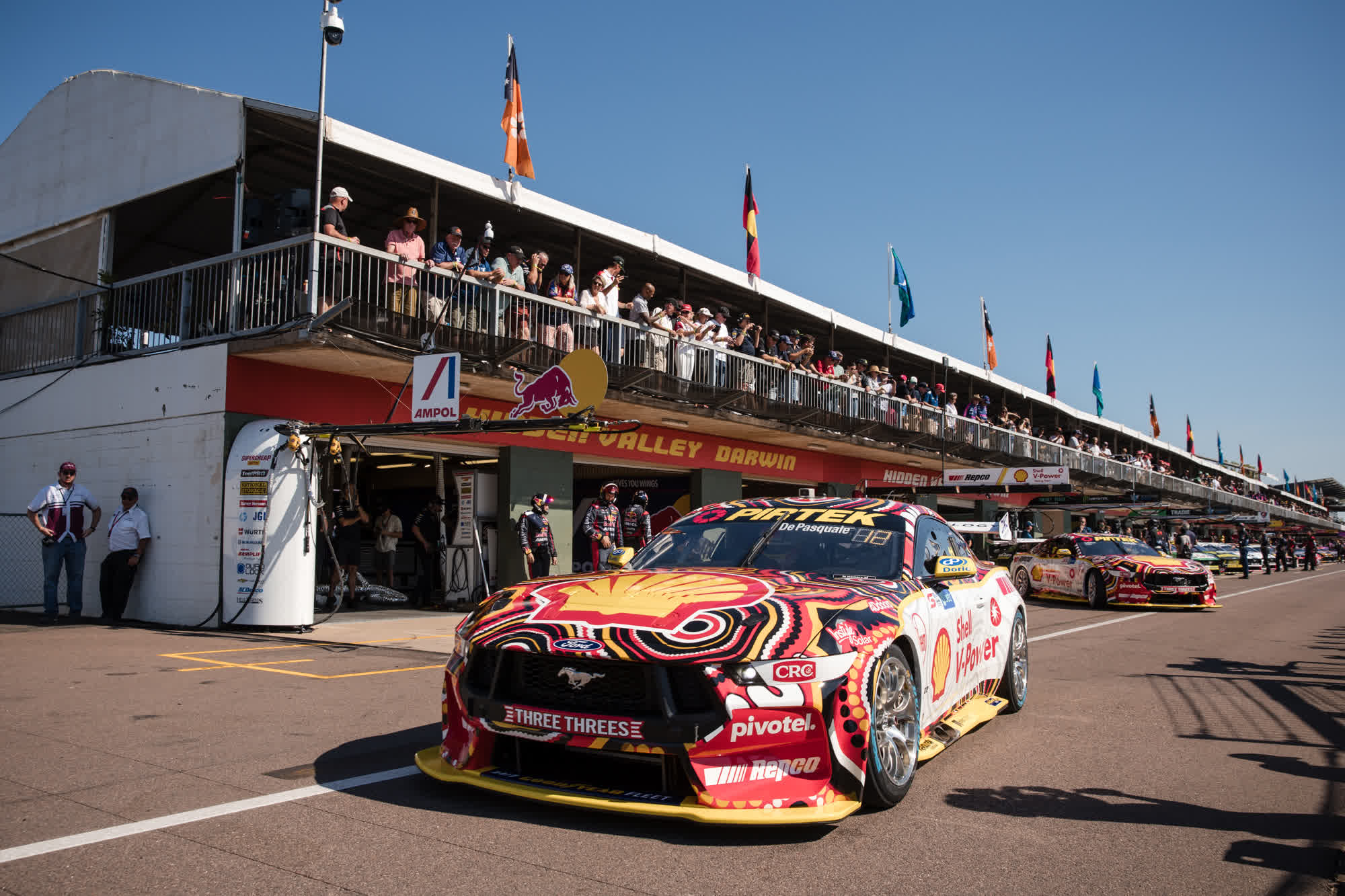 Race car with vibrant livery on a track, passing a pit stop area with spectators and flags. Sunny day at a motorsport event.