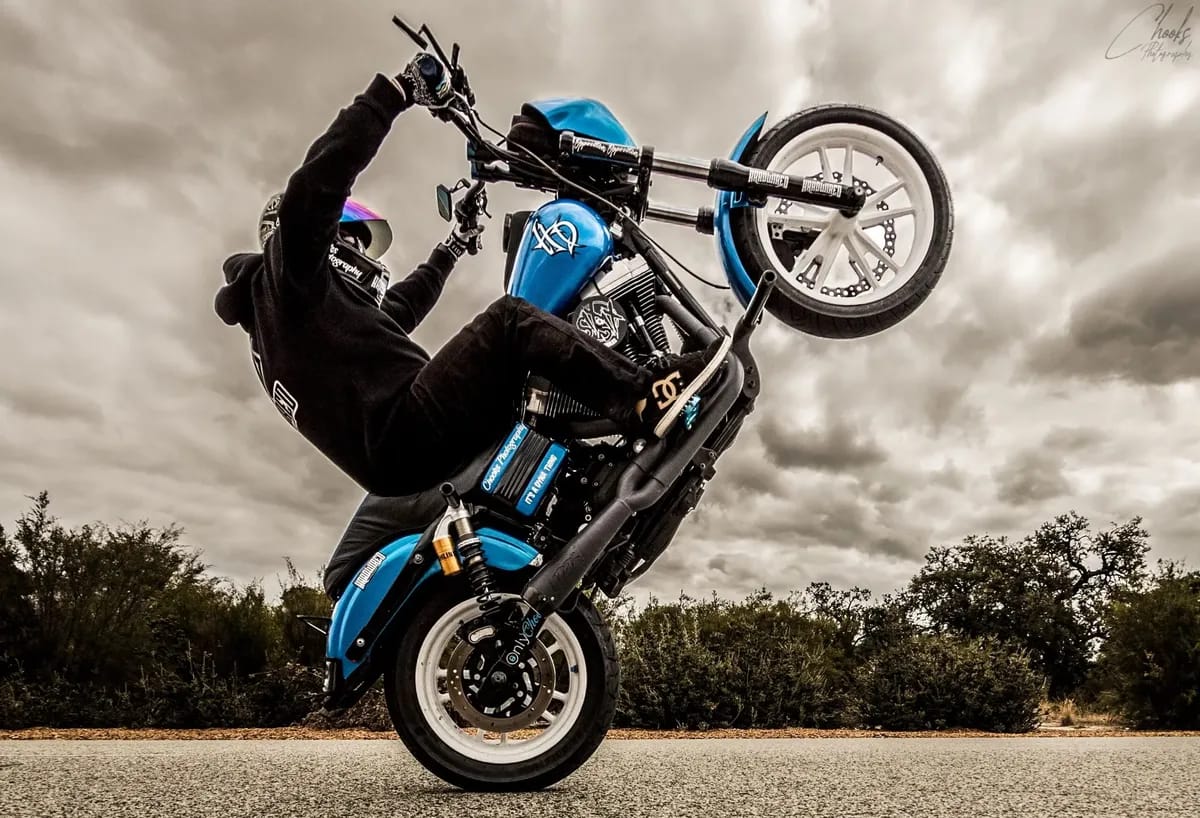 Motorcyclist in black gear performs a wheelie on a blue motorcycle on a cloudy day, with trees and a dramatic sky in the background.