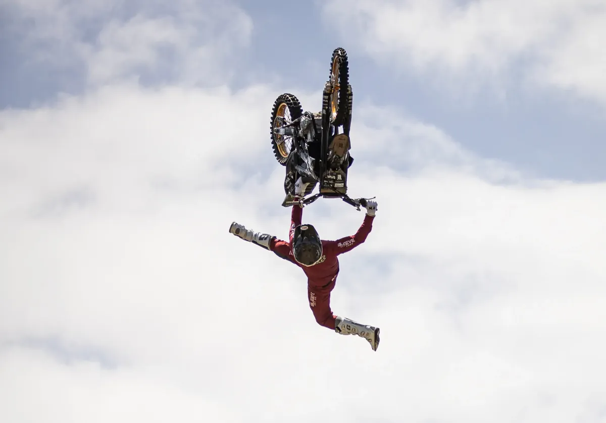 Motorcyclist in red suit performing an upside-down aerial stunt against a cloudy sky.
